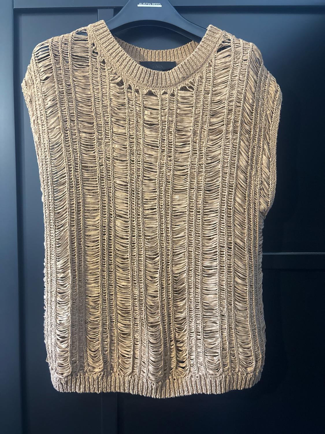 Beige sleeveless knitted top with an open-work and distressed design, hanging on a black hanger against a dark background. Text: AUSTIN REED, ZARA.
