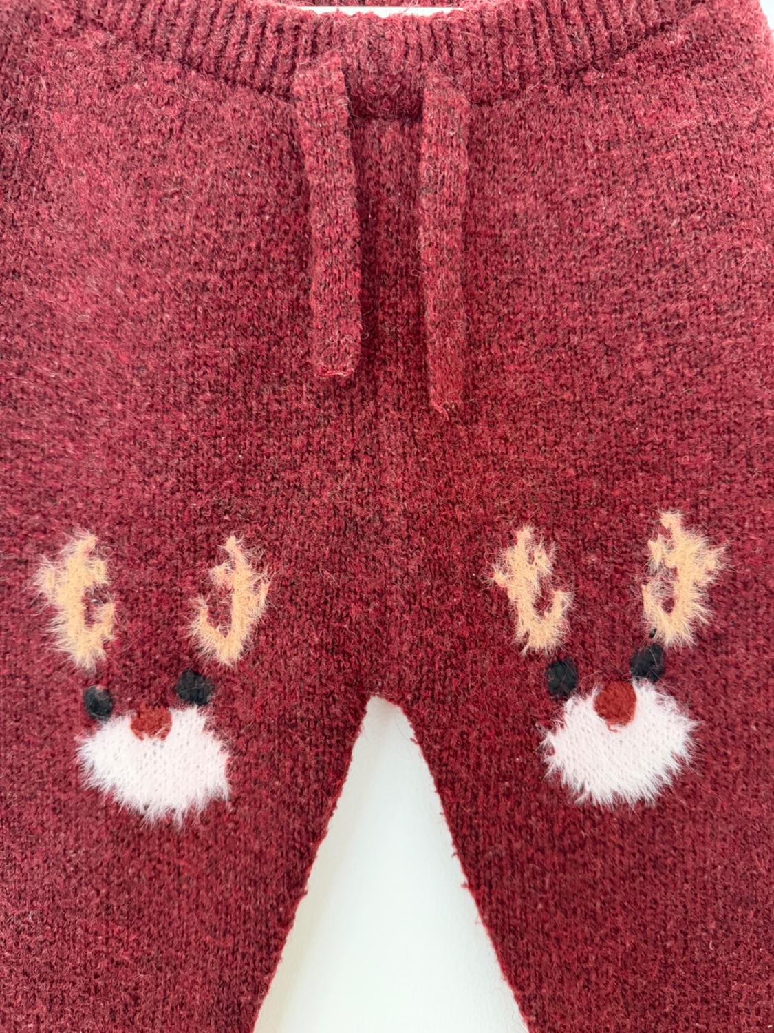 Close-up of maroon knitted trousers with a drawstring waist and two furry reindeer faces on the front. White background.