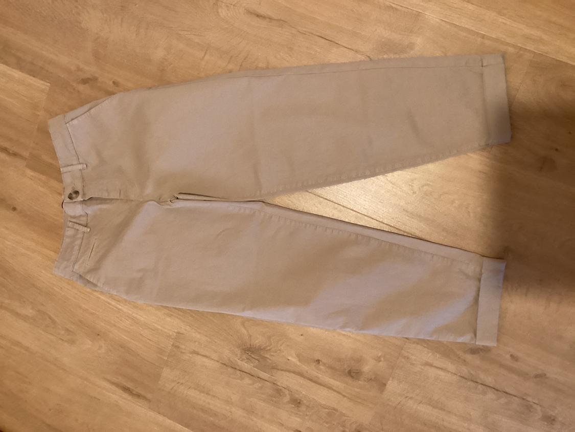 Beige straight-cut trousers on a light wooden floor, with button and zip fastening, and side pockets.