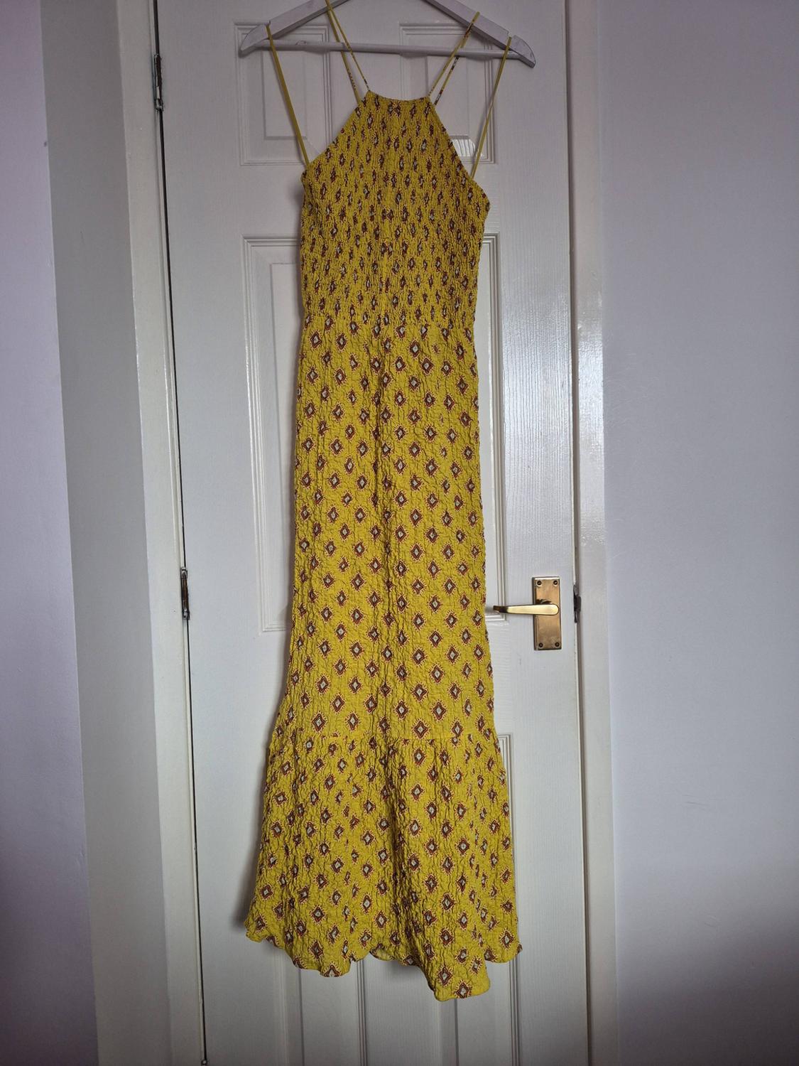 Long yellow dress with a diamond print, halter neckline and ruched bodice, hanging on a hanger on a white door.