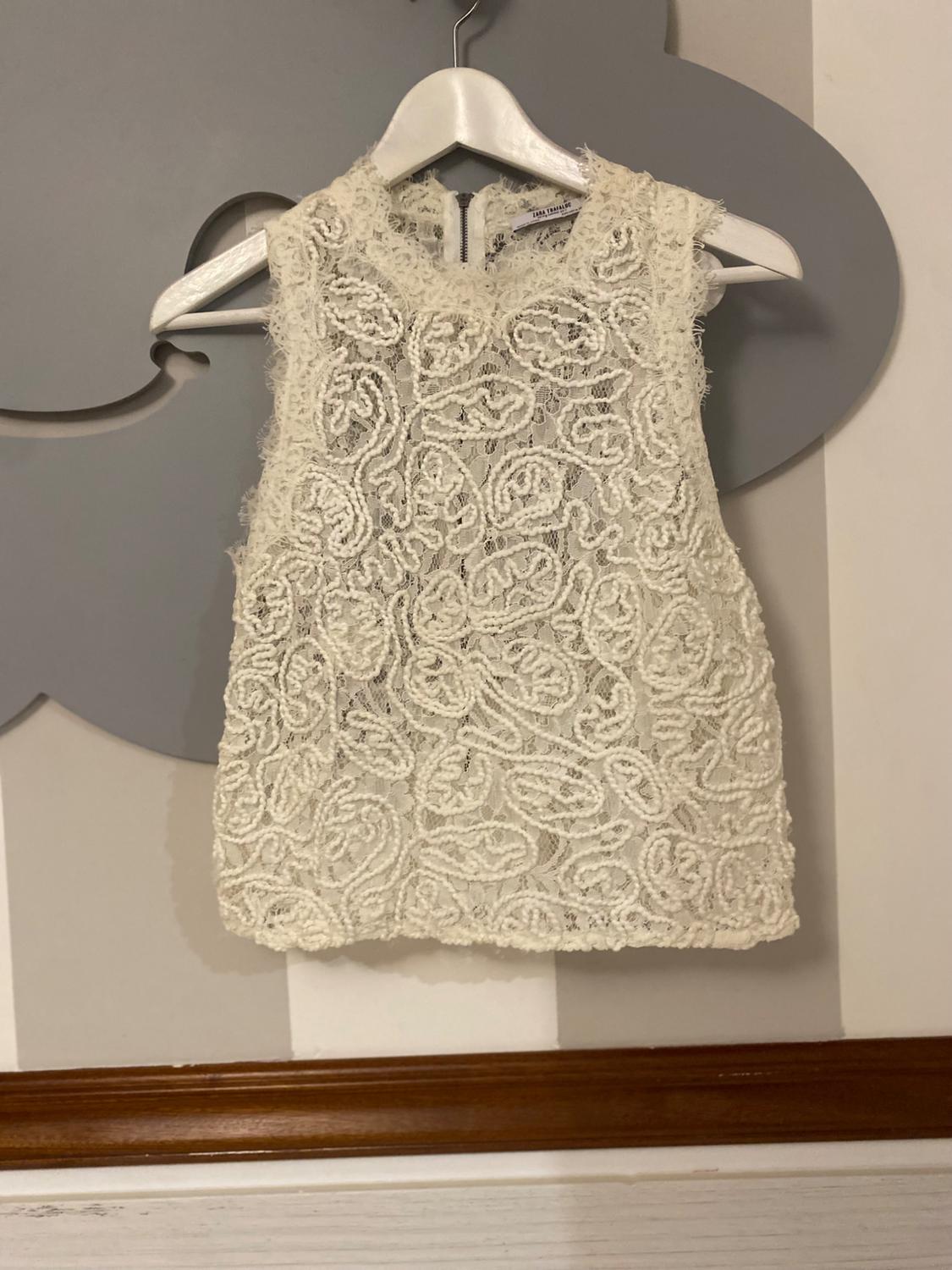 White sleeveless textured lace top on a white hanger against a striped wall with a grey cloud decoration. ZARA TRAFALUC label.