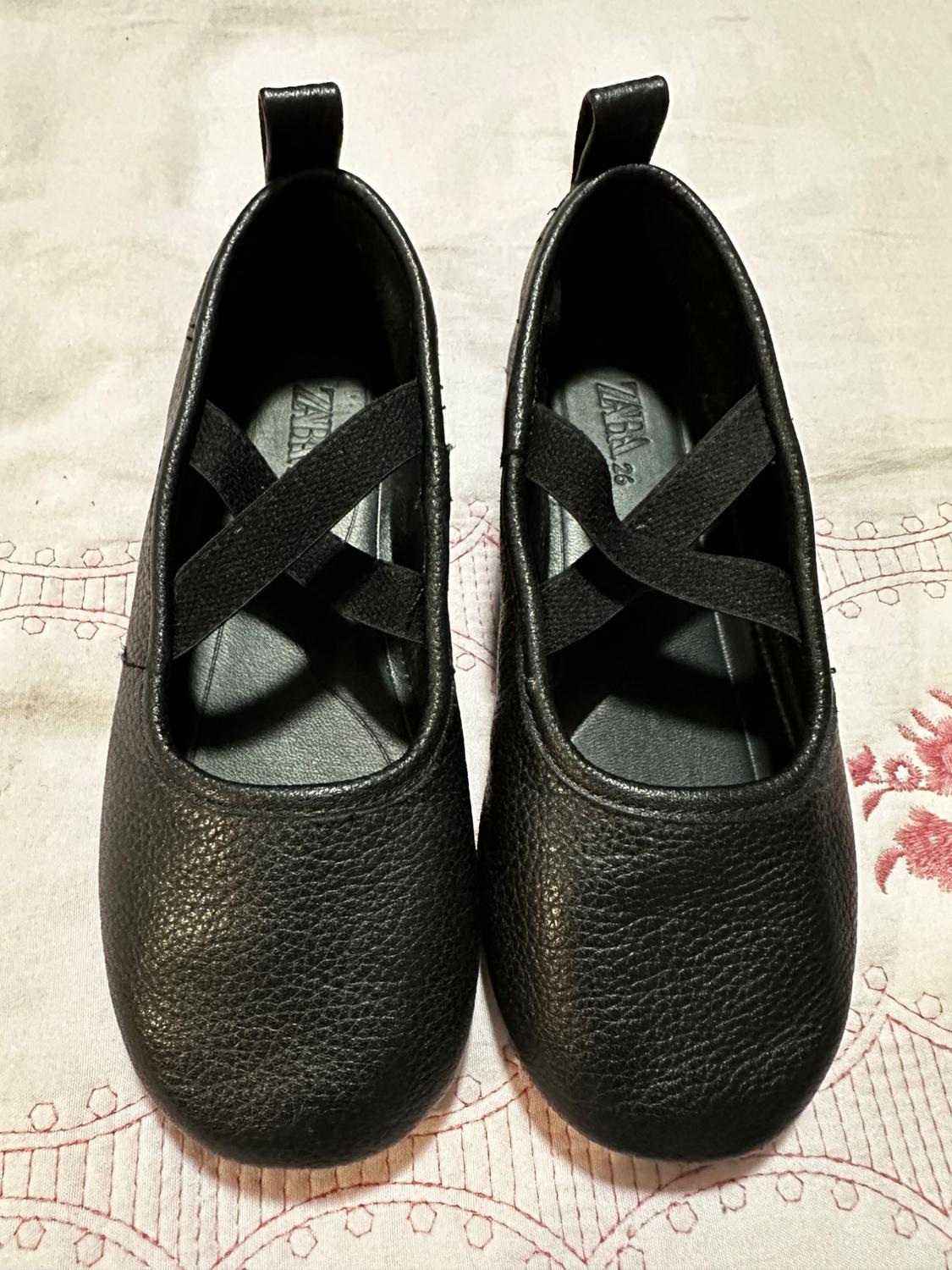 Black round-toe shoes with crossover straps, on a fabric with red embroidery. Visible text: "ZARA 36".