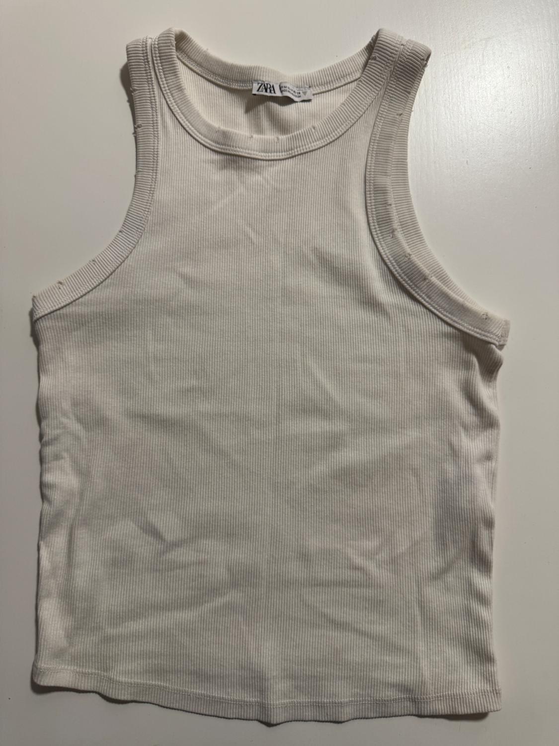 White ribbed vest top with a round neckline and raw edges on a white background. Label with the text ZARA.