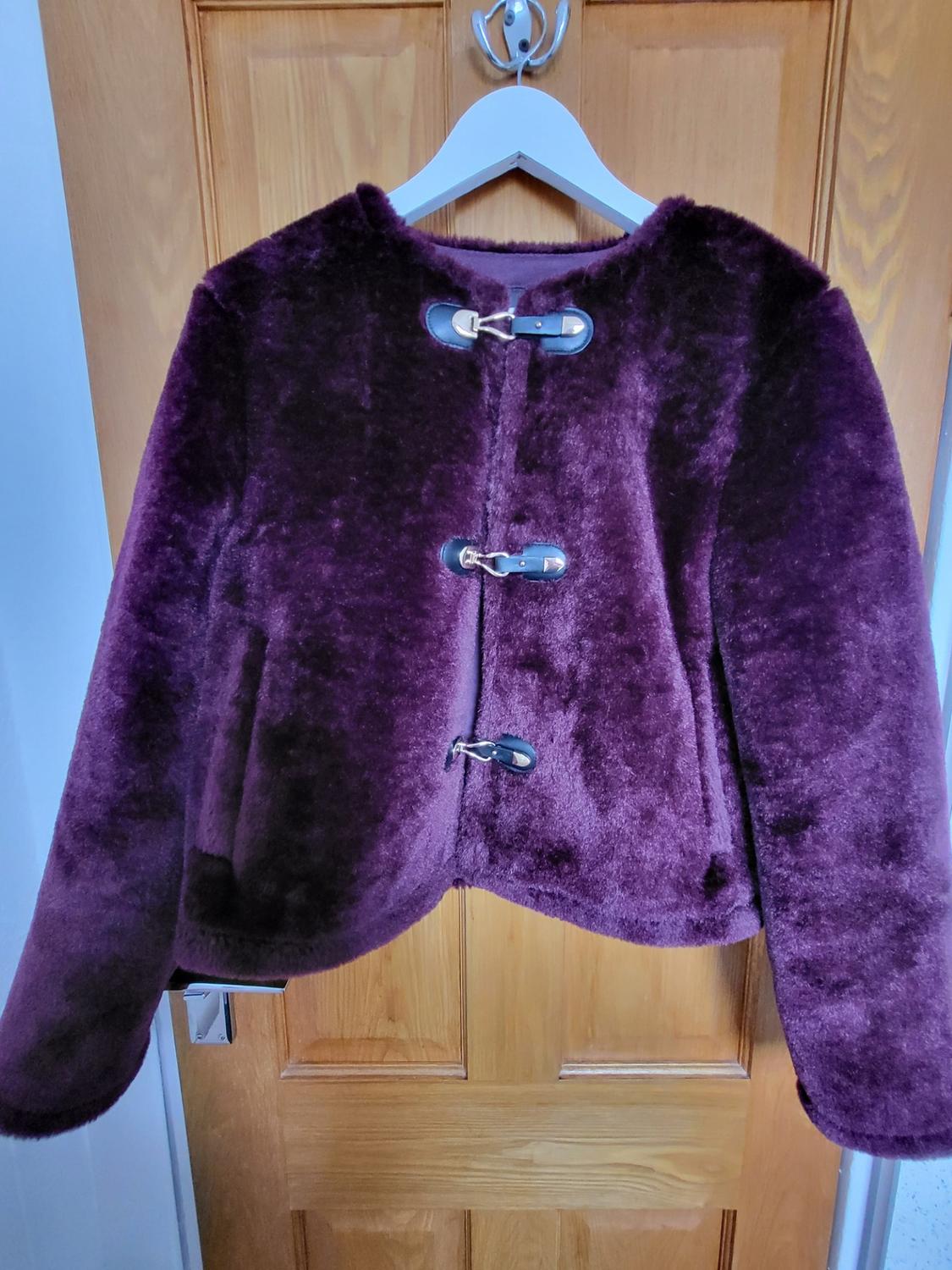 Dark plum faux fur jacket with three front fastenings, hanging on a white hanger on a wooden door.