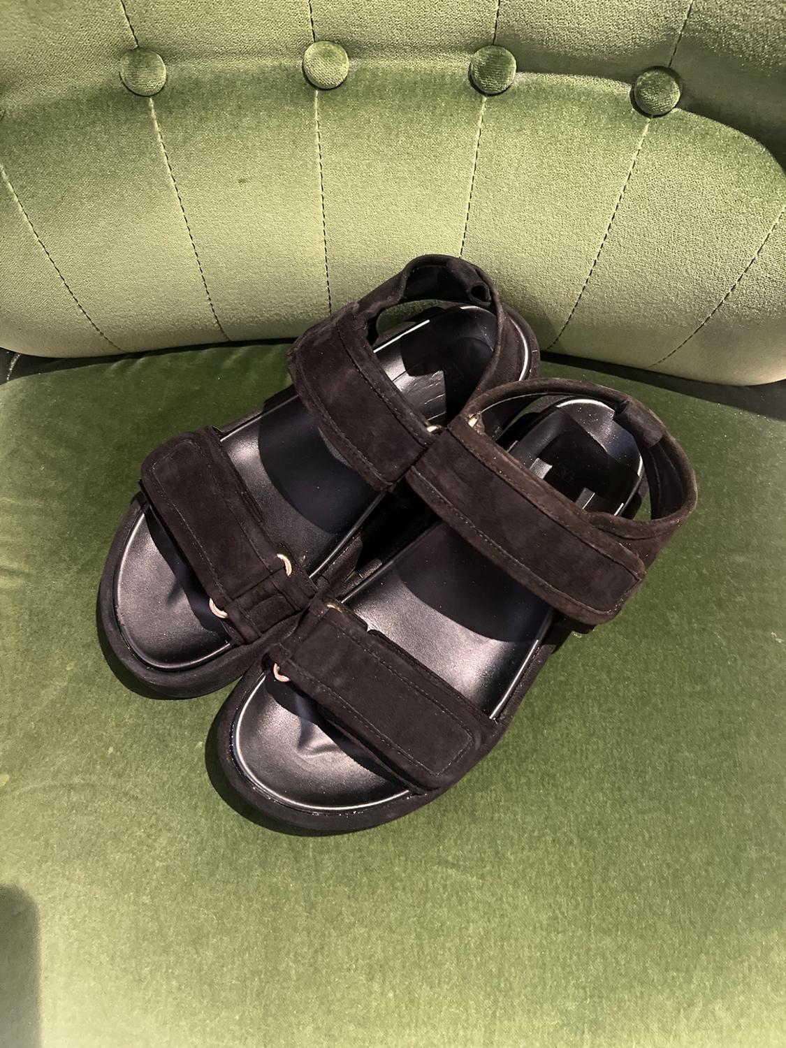 Pair of black sandals with wide straps and a chunky sole, displayed on a green velvet armchair.