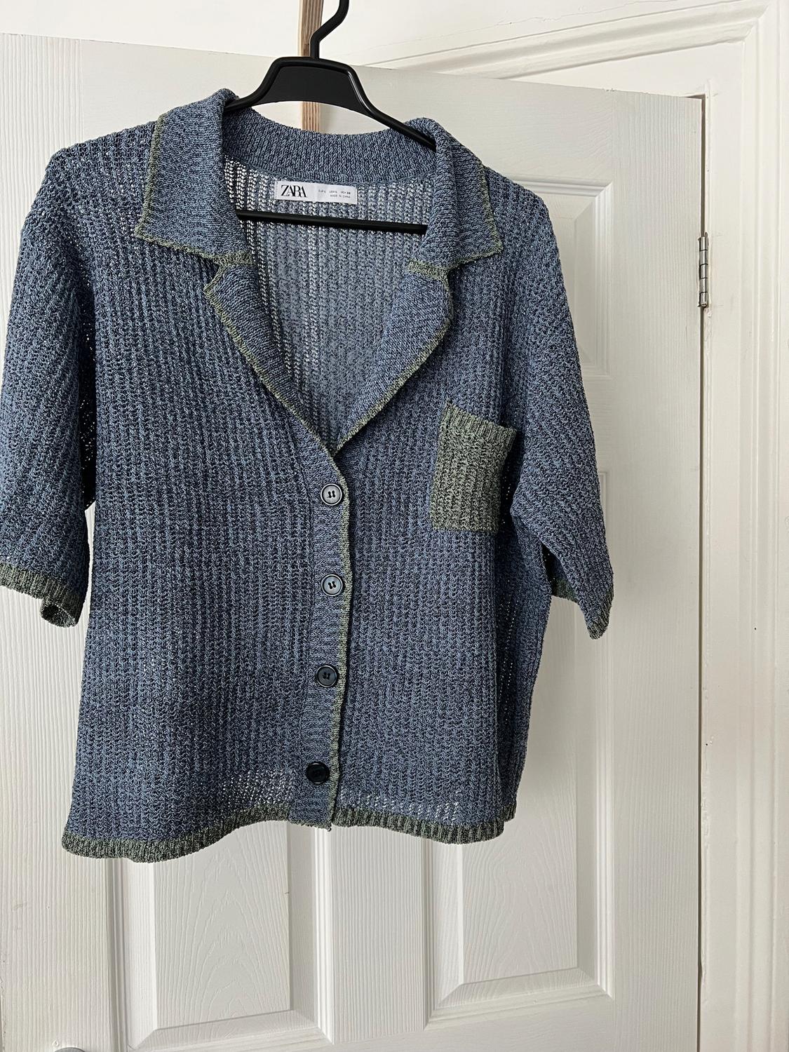 Blue knitted cardigan with buttons and a chest pocket, hanging on a black hanger in front of a white door.