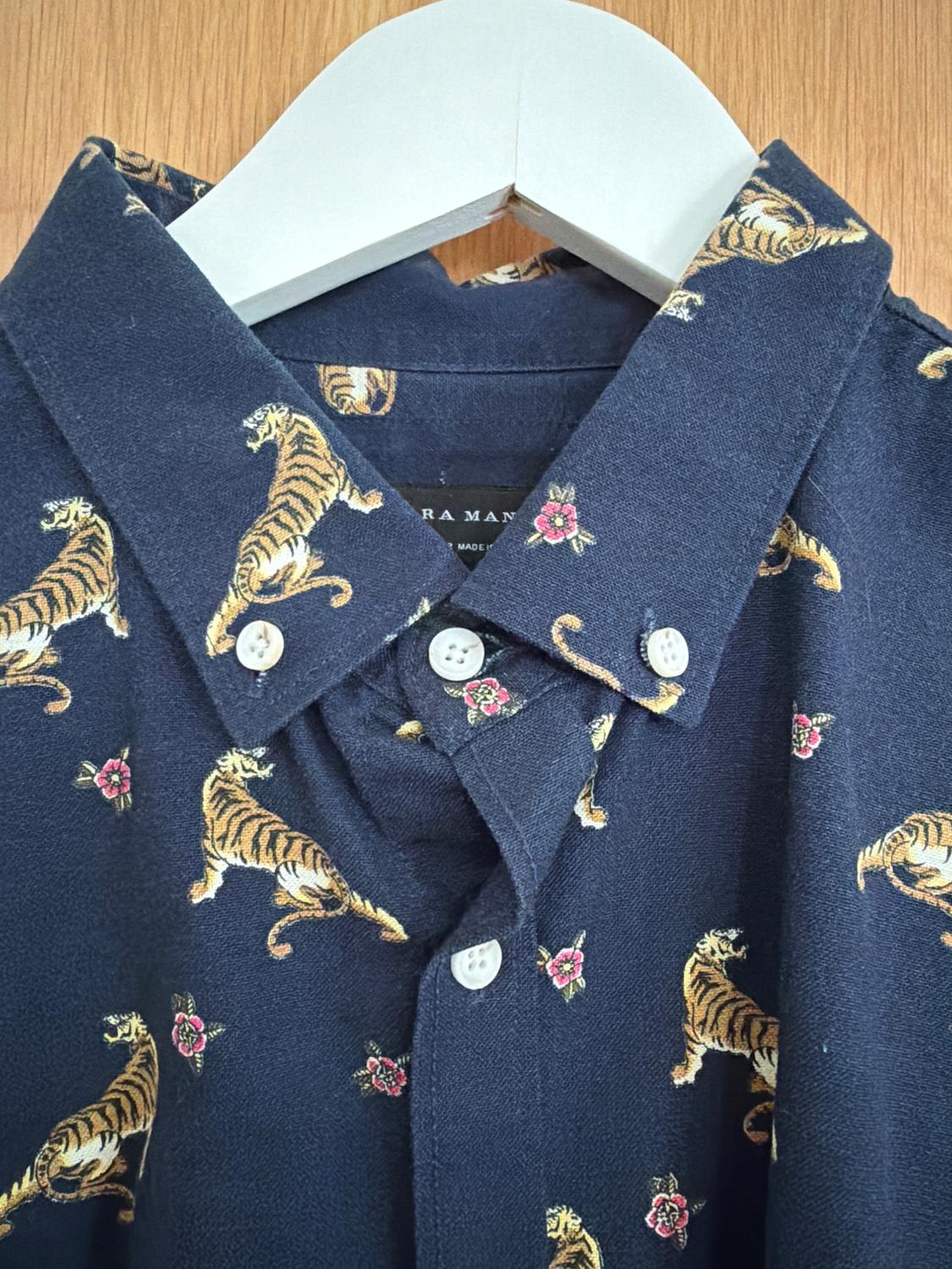 Blue shirt with tiger and pink floral print, hanging on a white hanger against a wooden background.
