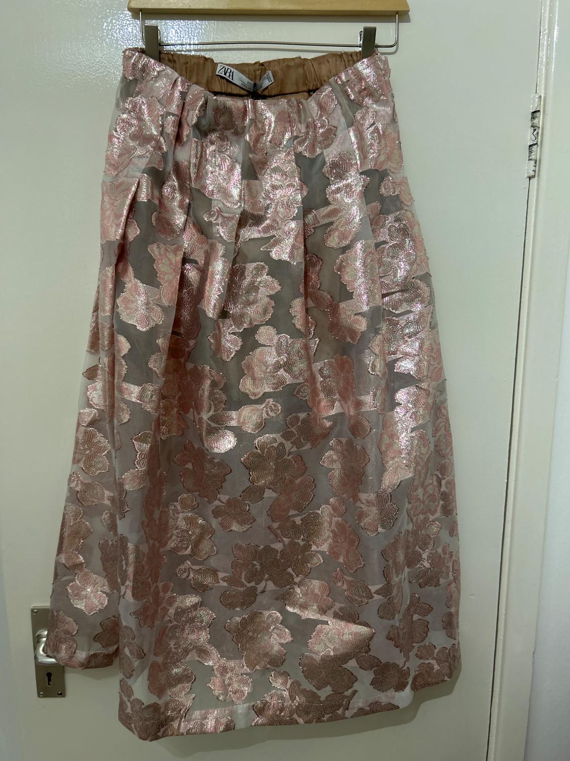 Skirt hanging on a hanger, with a floral print in pink and gold tones on a sheer background. Visible label at the waist.