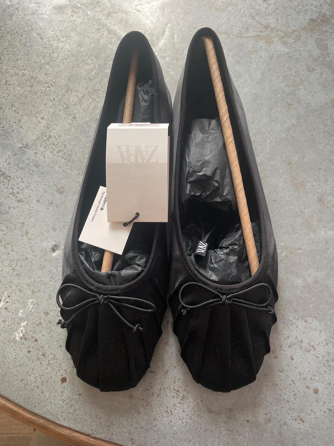 Black flat shoes with a bow, stuffed with paper, on a grey table. Visible label with the text "ZARA".