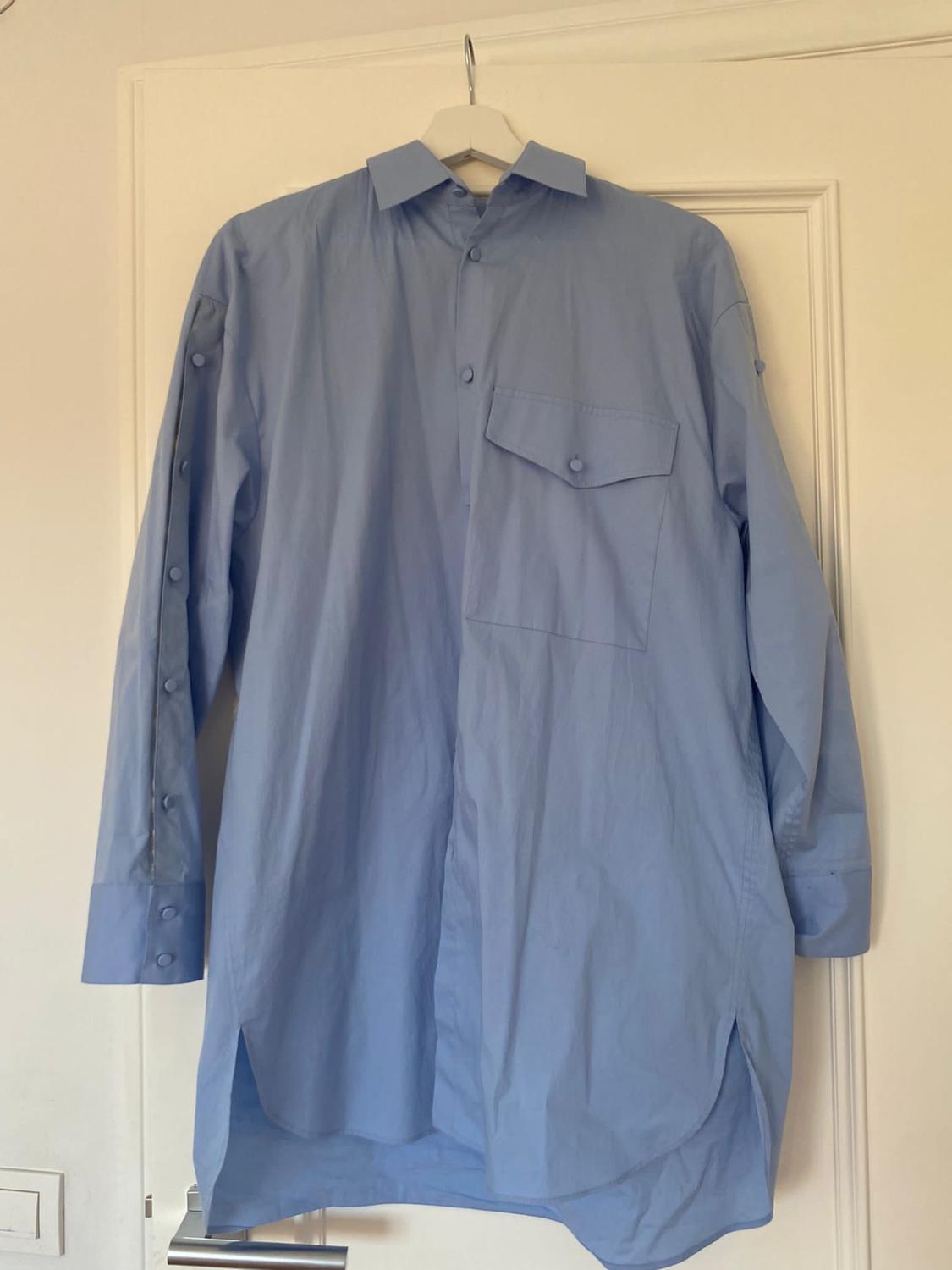 Light blue, long-sleeved shirt hanging on a white hanger over a white door. Chest pocket and buttons on the cuffs.