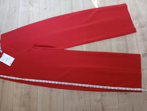 Red trousers laid out on a light wooden floor, with a white tape measure along one leg and a visible label.