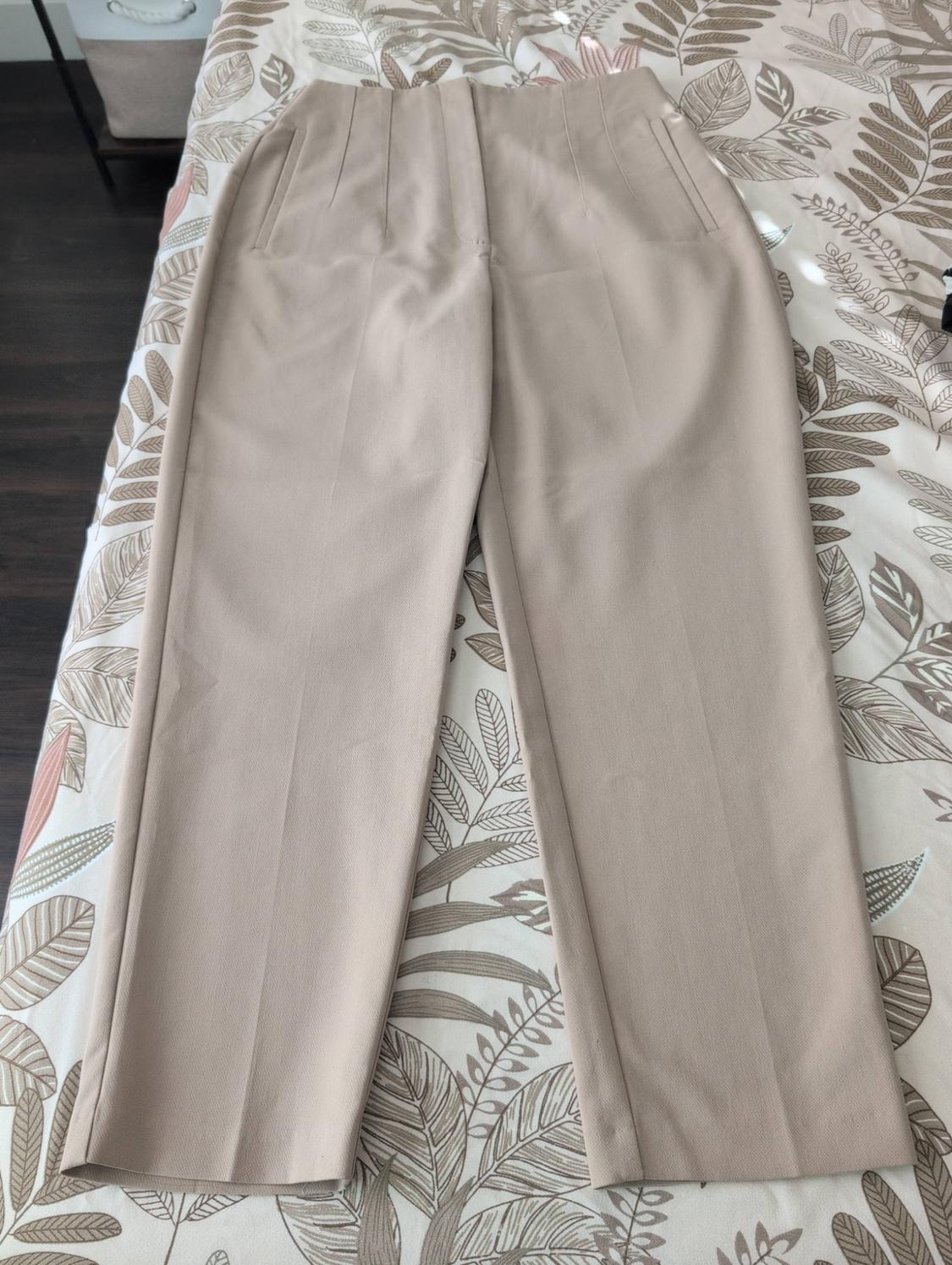 Beige pleated dress pants, laid out on a bed with leaf-patterned sheets.