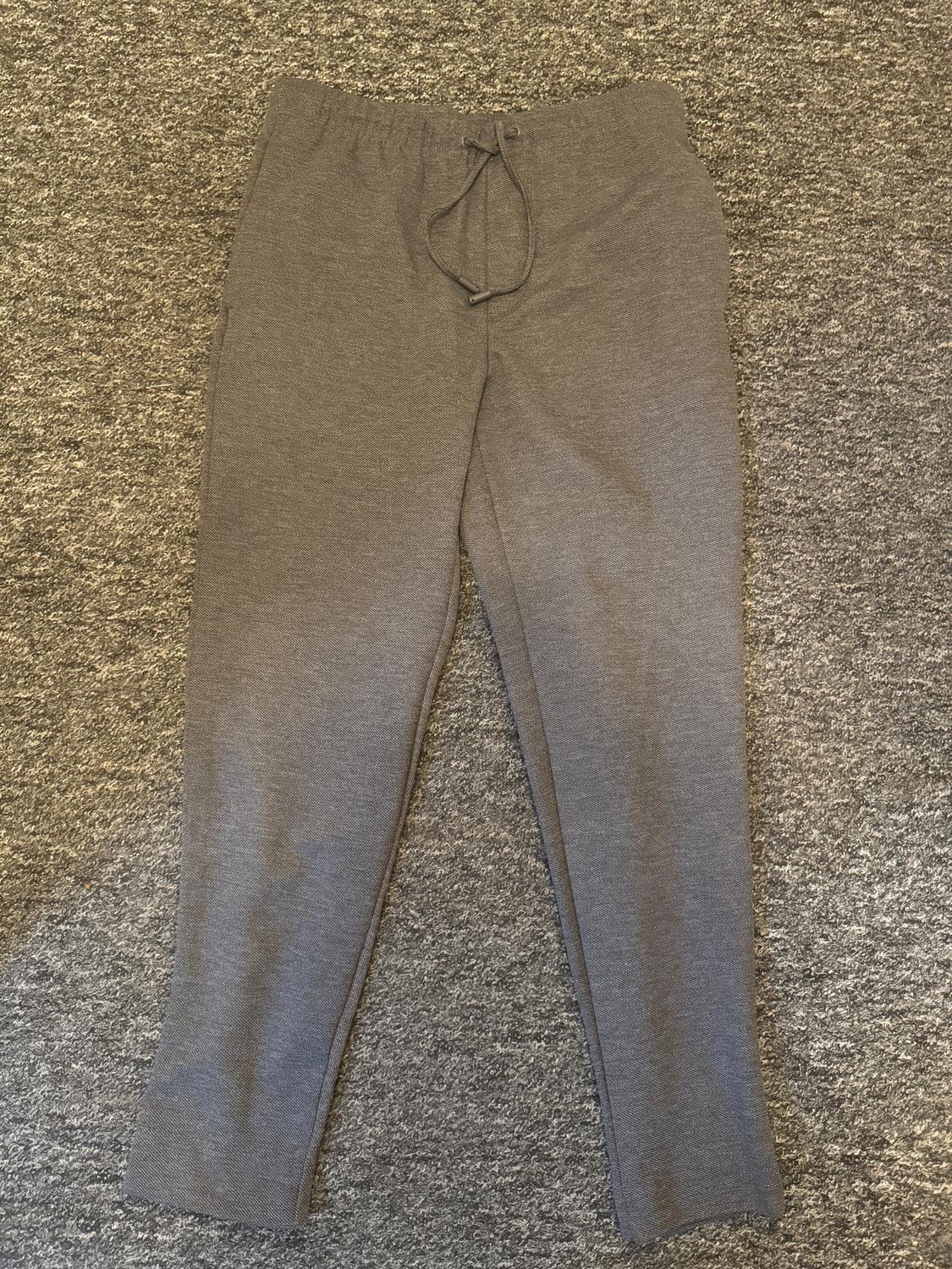 Grey knitted trousers with an elasticated drawstring waist, placed on a similar textured grey rug.