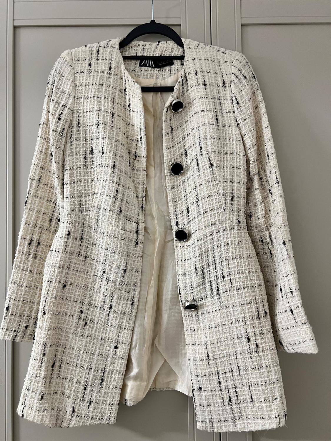 White textured fabric coat with black buttons, hanging on a black hanger. Visible label with the text "ZARA".