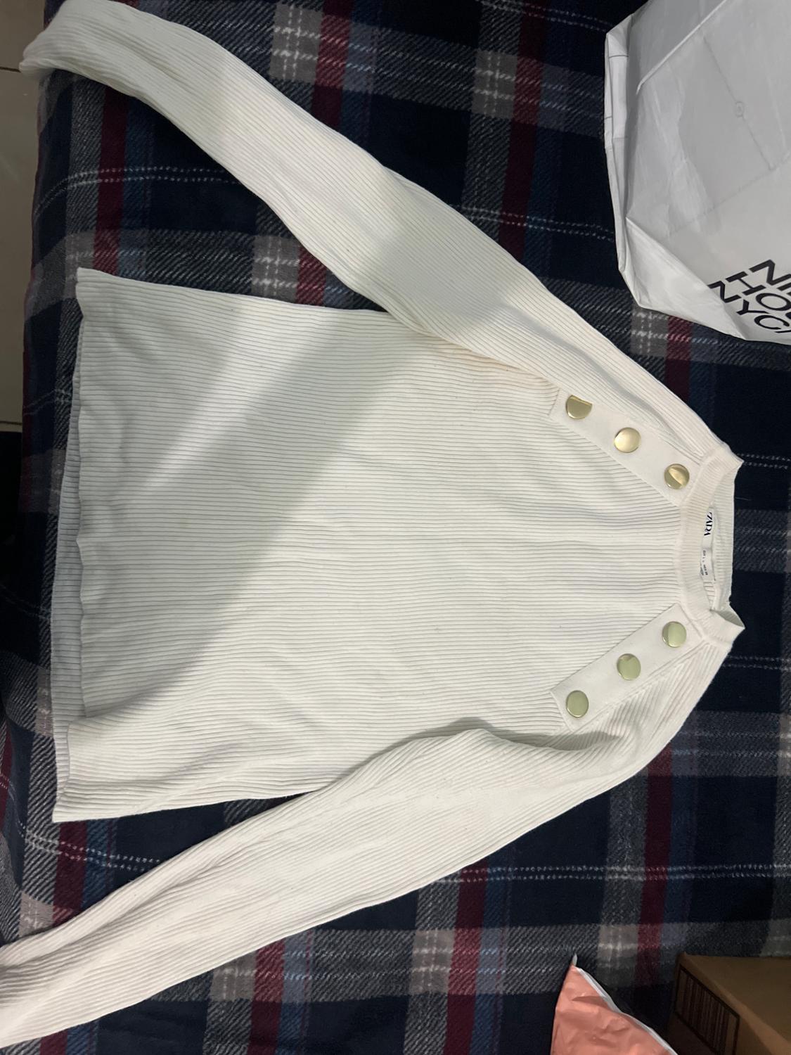 White long-sleeved jumper with gold buttons on the shoulder, over a dark tartan blanket. White bag to the right.