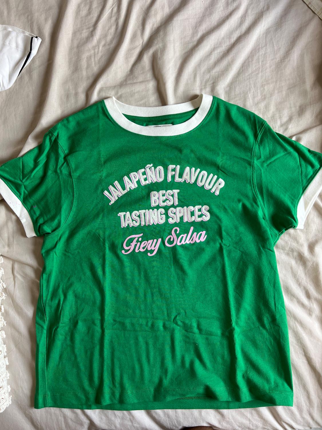 Green t-shirt with white trim on a beige bed. Text: "Jalapeño Flavour Best Tasting Spices Fiery Salsa".