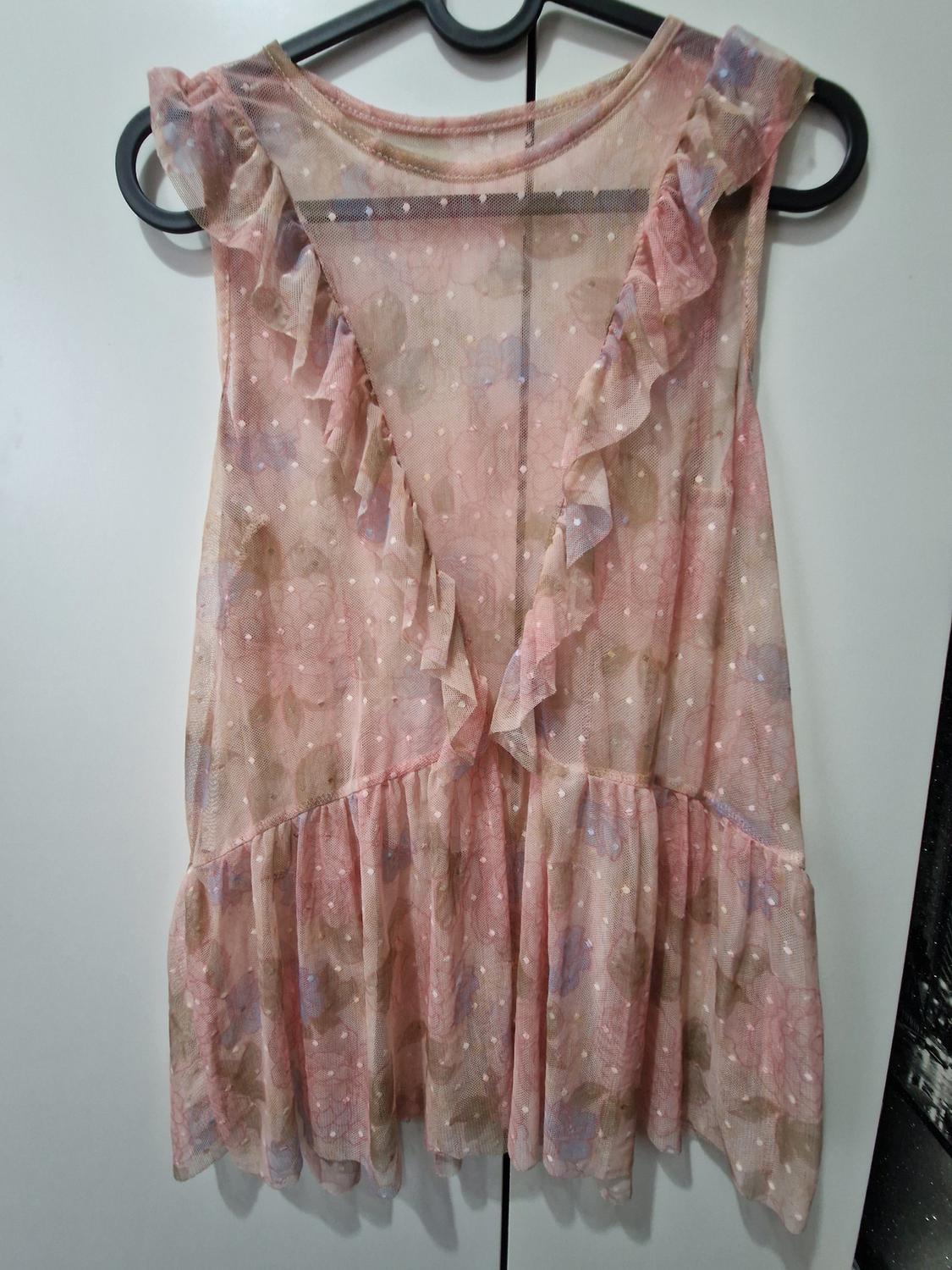 Pink semi-sheer blouse with a floral and polka dot print. Sleeveless, with a V-neck, frills and a gathered hem, hanging on a hanger.
