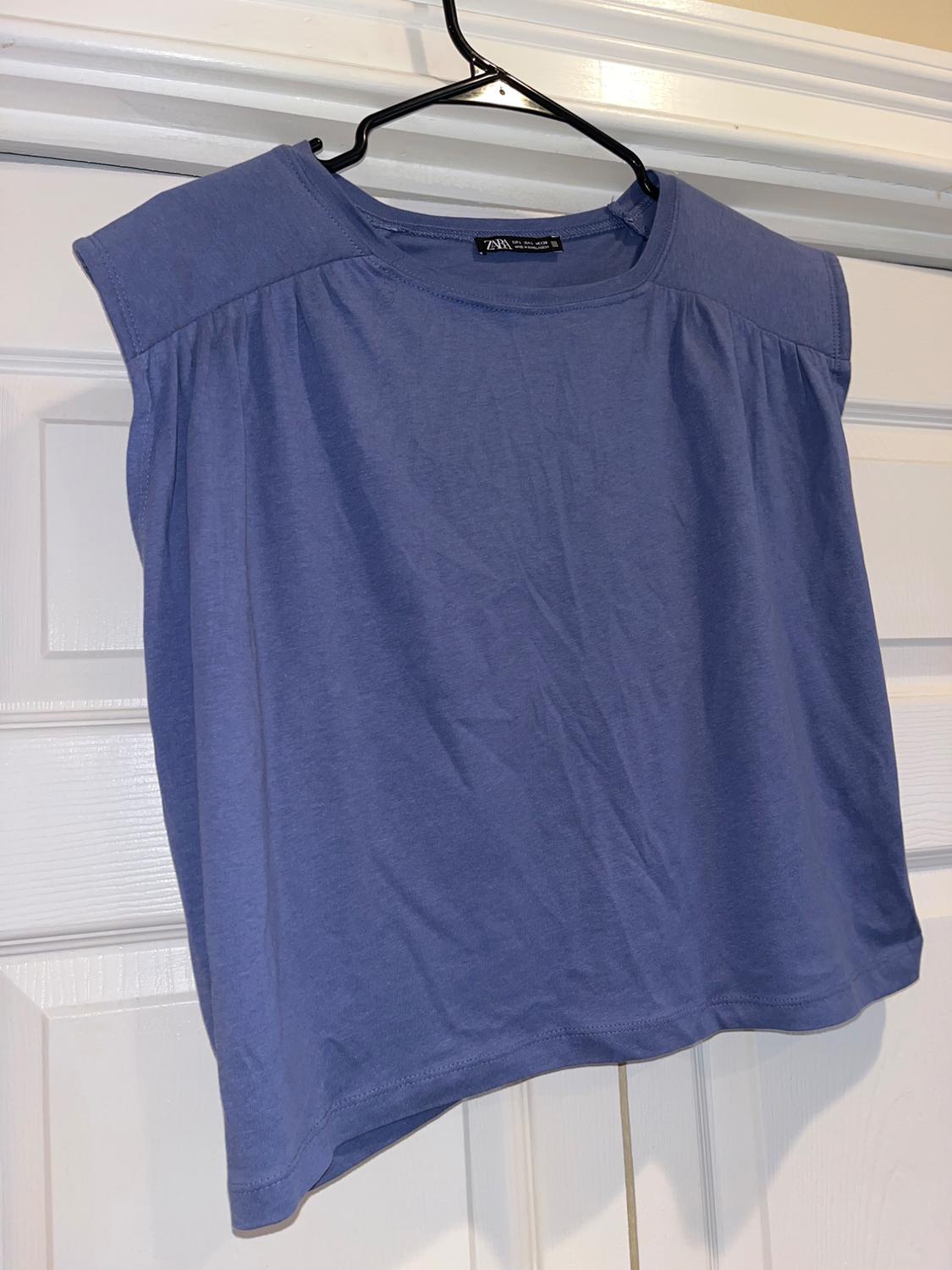 Blue sleeveless top hanging on a black hanger over a white door. Visible label on the collar.