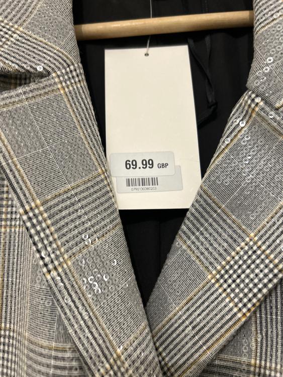 Price tag for £69.99 on a grey checked coat with sequins, hanging on a wooden hanger.