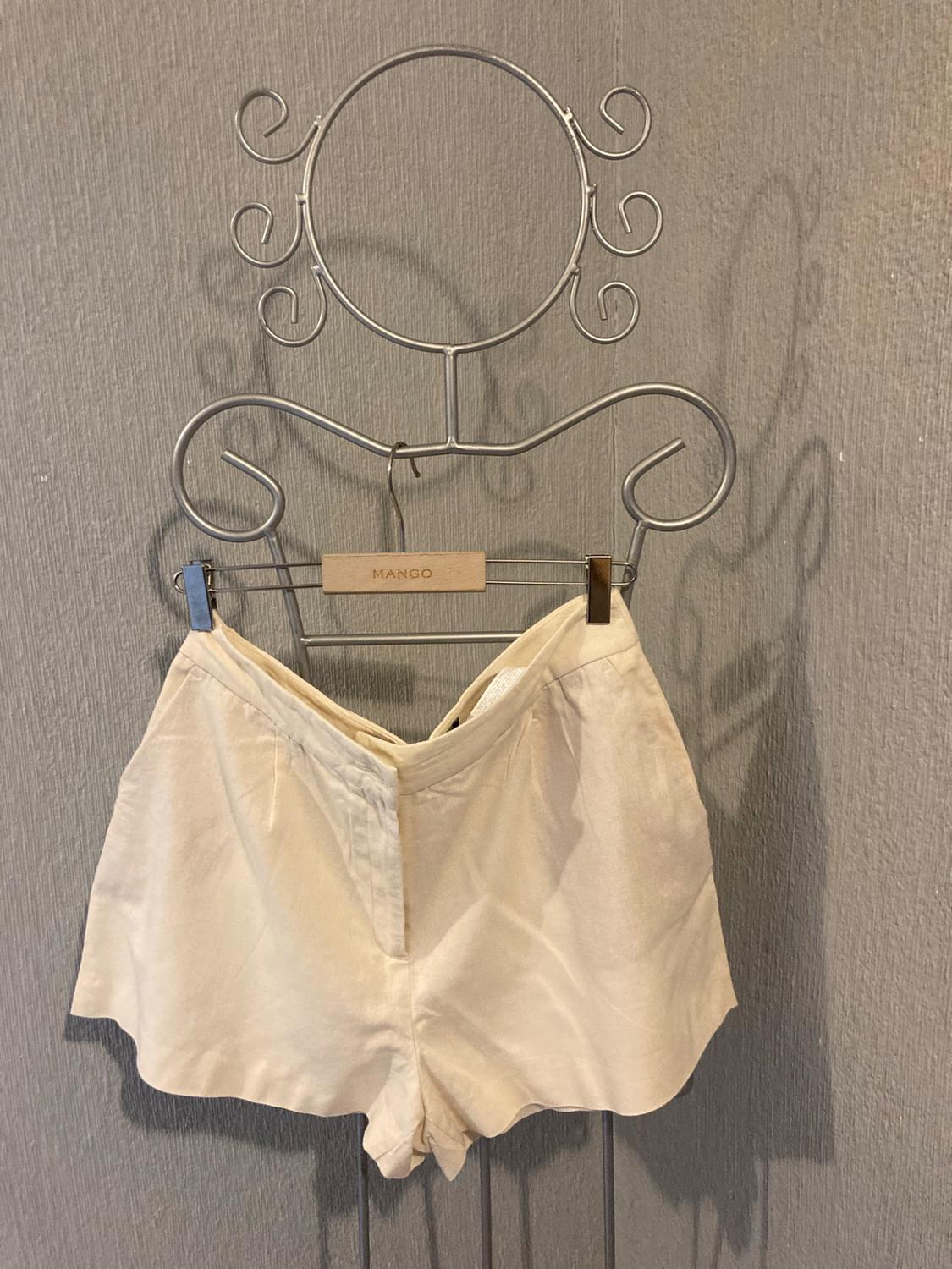 Beige shorts on a hanger with MANGO text, hanging on a decorative wire rack in front of a grey wall with shadows.