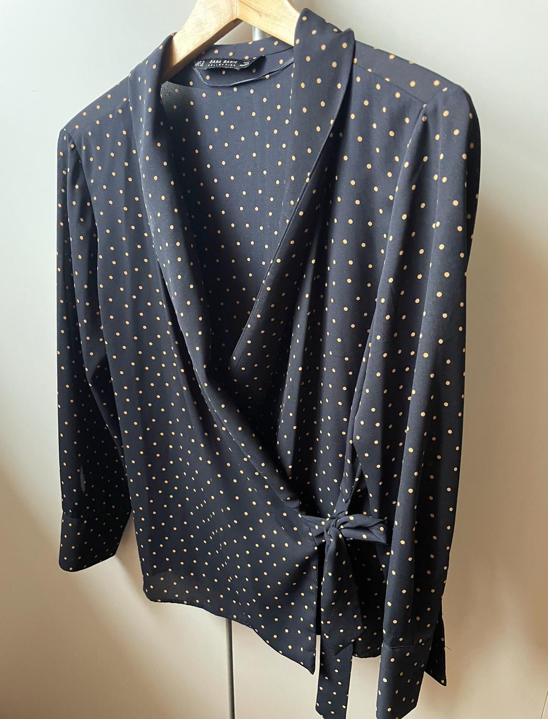 Front view of a black blouse with gold polka dots and a side bow, long sleeves.