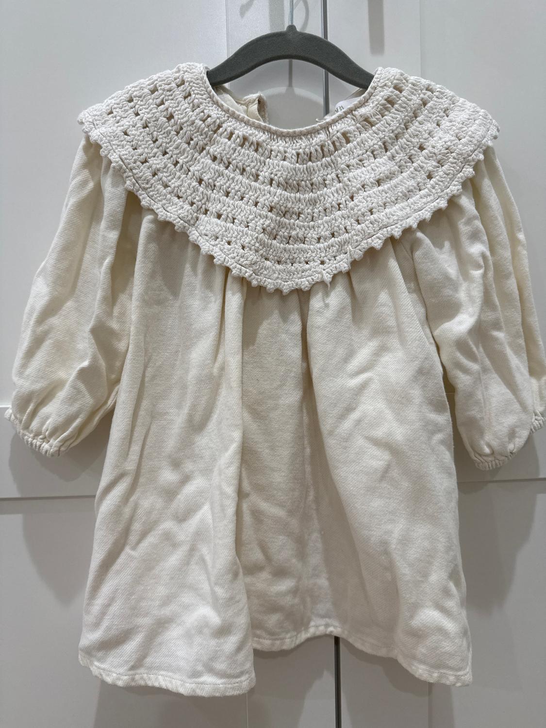 Cream children's dress with crochet knit collar, puffed long sleeves, hanging on a black hanger against a white background.