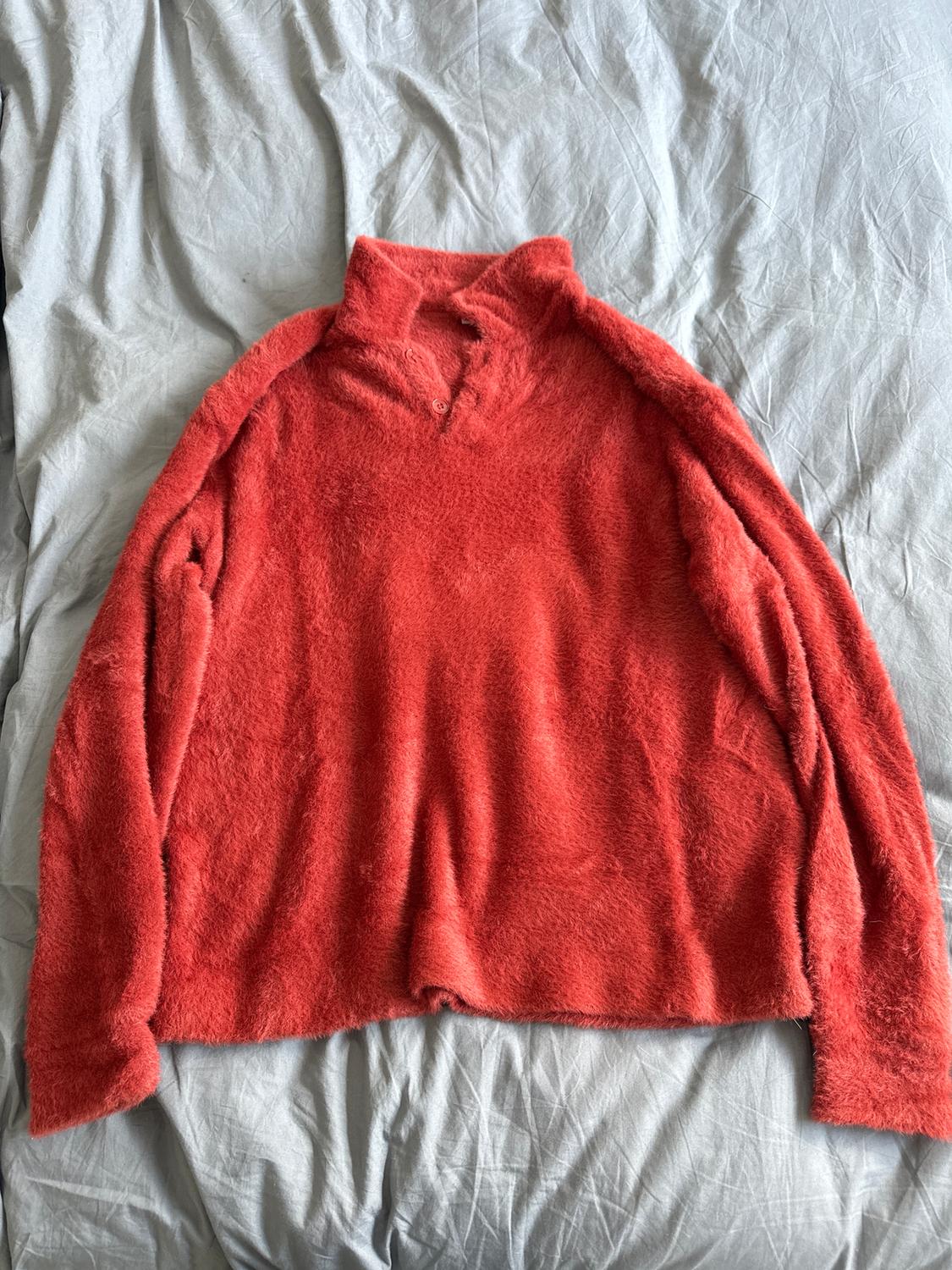 Red fleece jumper with a high neck and buttons, spread out on a bed with wrinkled grey sheets.