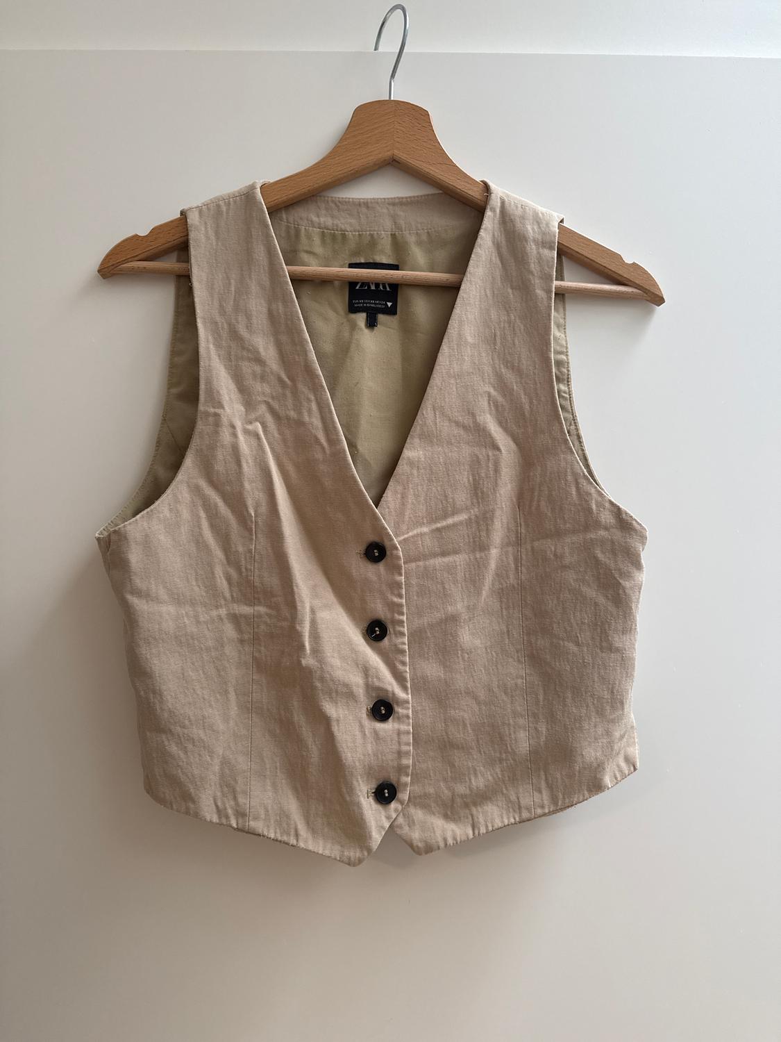 Beige sleeveless waistcoat with black buttons, hanging on a wooden hanger. Label visible inside. White background.
