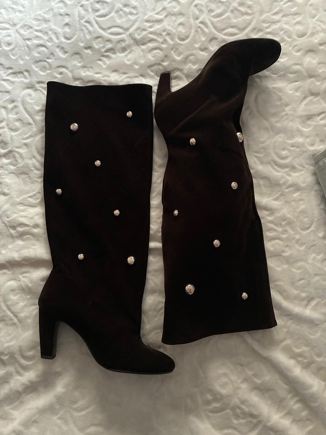 Black high-heeled boots with a chunky heel and pearl embellishments, placed on a white embossed bedspread.