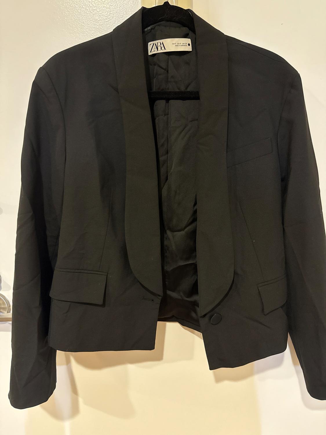 Women's black jacket hanging on a hanger, with wide lapels and front pockets. Visible label: ZARA. White background.