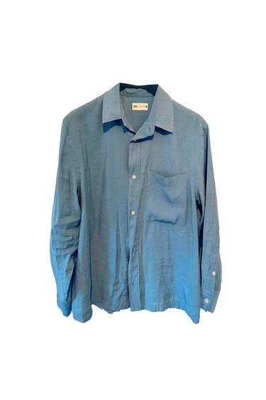 Blue, long-sleeved shirt with a chest pocket and white buttons, hanging against a white background. Label visible on the collar.