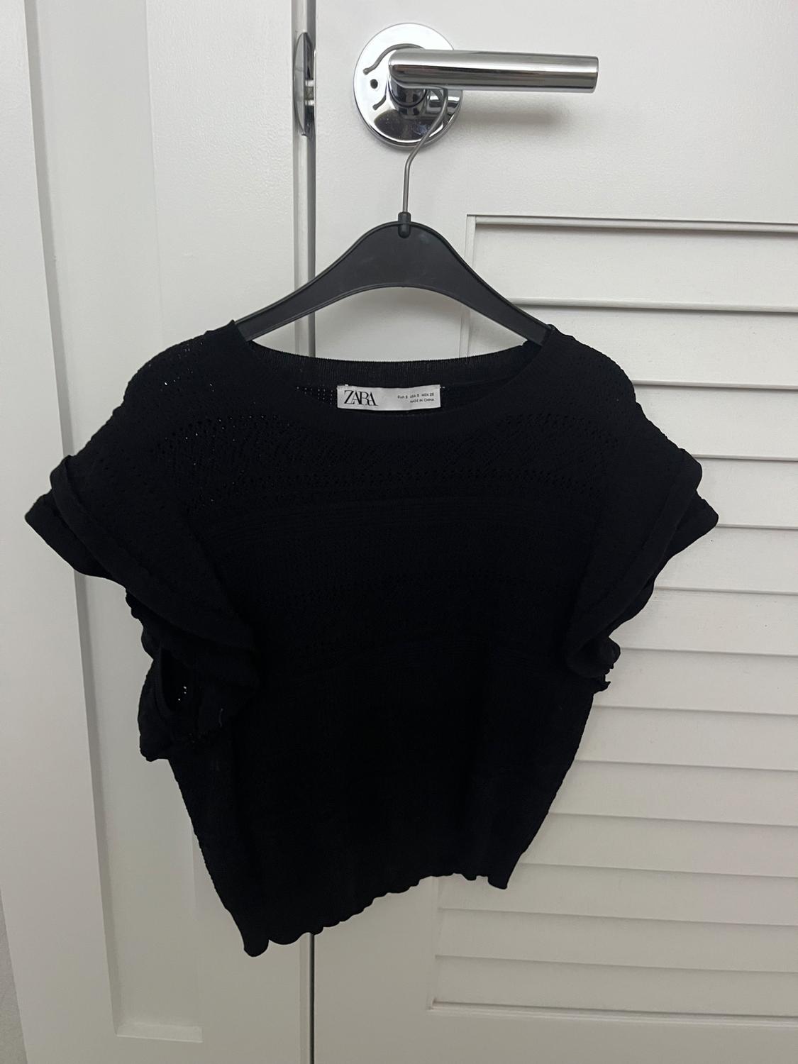 Black knitted jumper hanging on a black hanger over a white door. Visible label: "Zara".