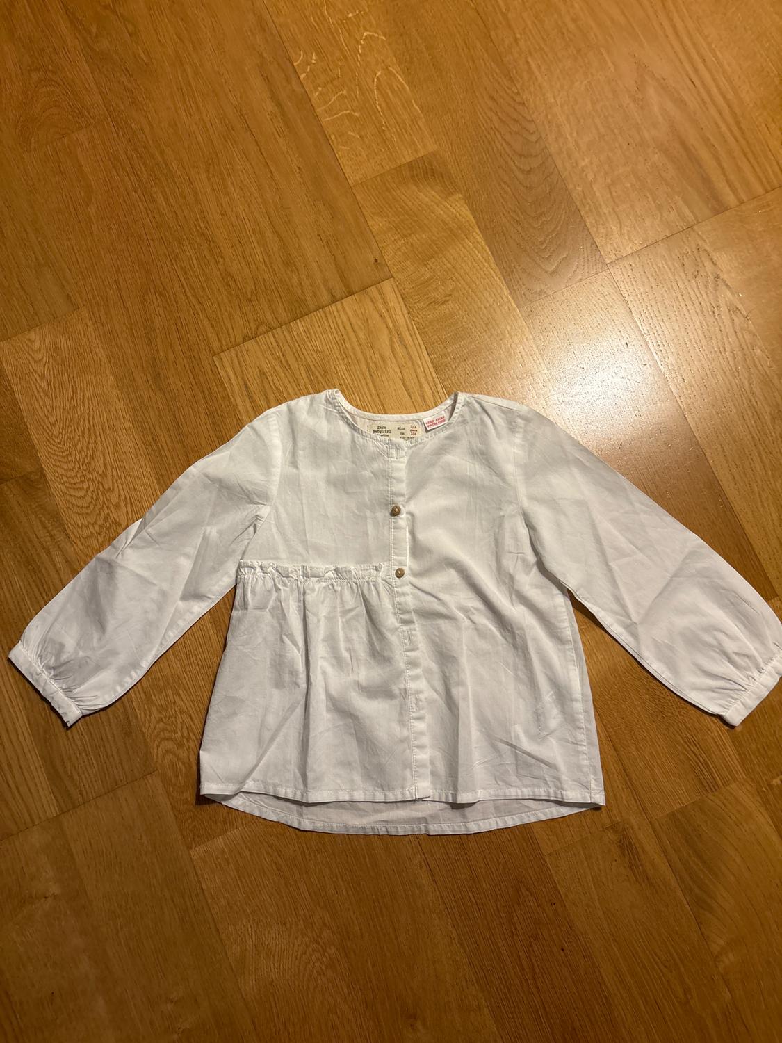 White long-sleeved button-up blouse, laid on a light wooden floor.