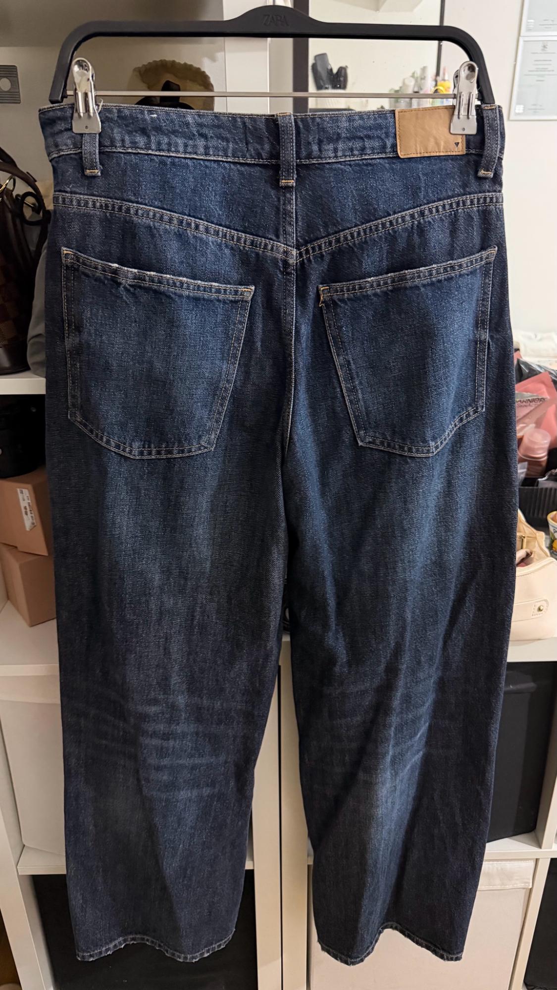 Rear view of dark blue jeans on a black hanger with the ZARA label. Their pockets and brown patch stand out against a white shelf.