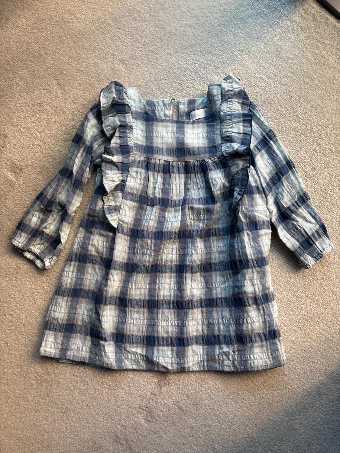 Blue and white checked blouse with ruffles on the sleeves and chest, on a beige rug illuminated by natural light.