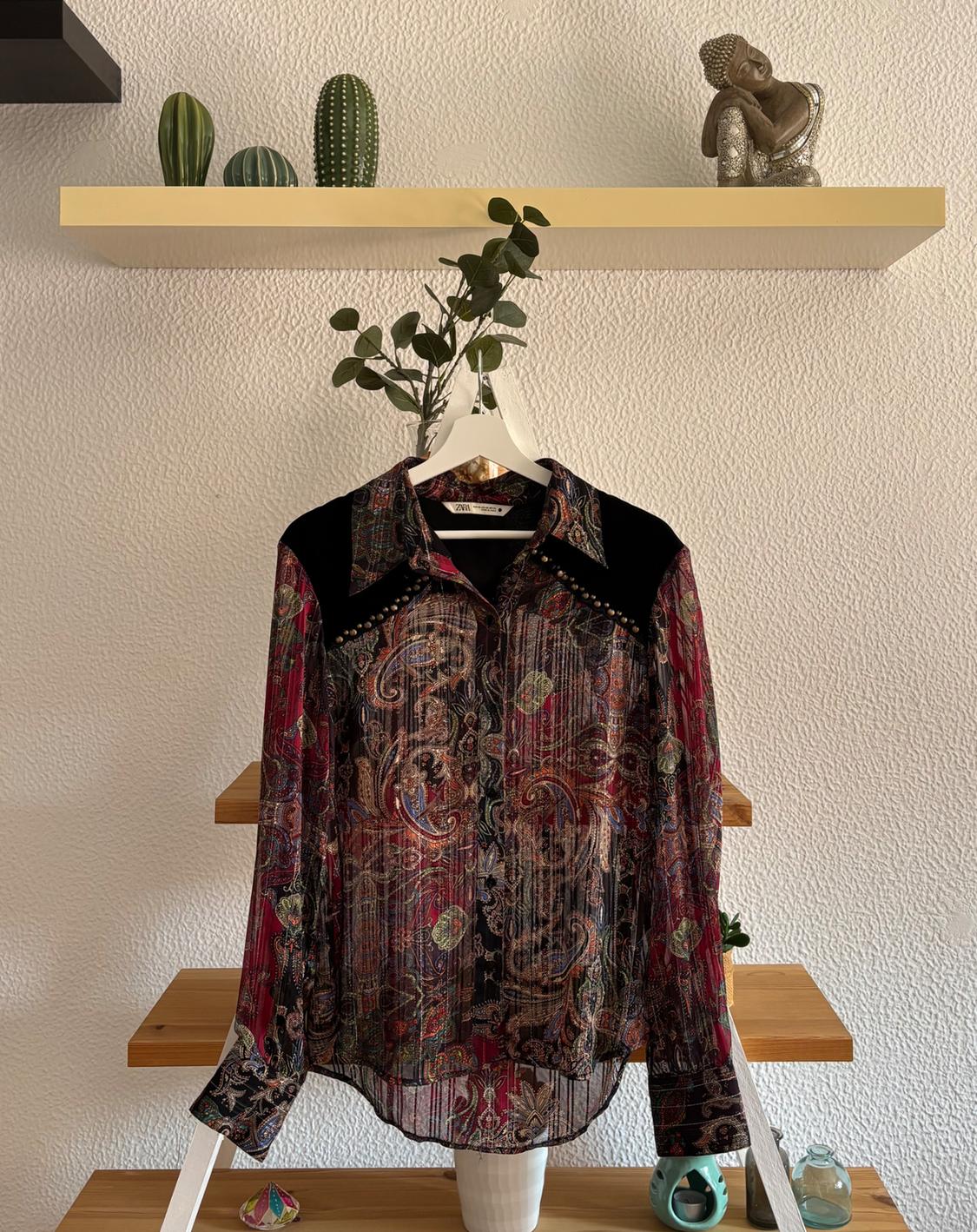 Coloured patterned shirt hanging on a white hanger over a wooden ladder. Background with shelf, cactus, and decorative figure.