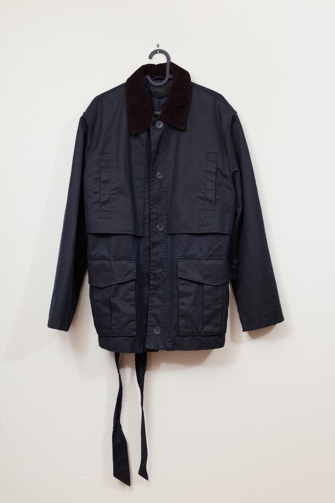 Navy blue utility-style jacket with a dark corduroy collar, front pockets with a flap, and a dangling belt, hanging on a white background.