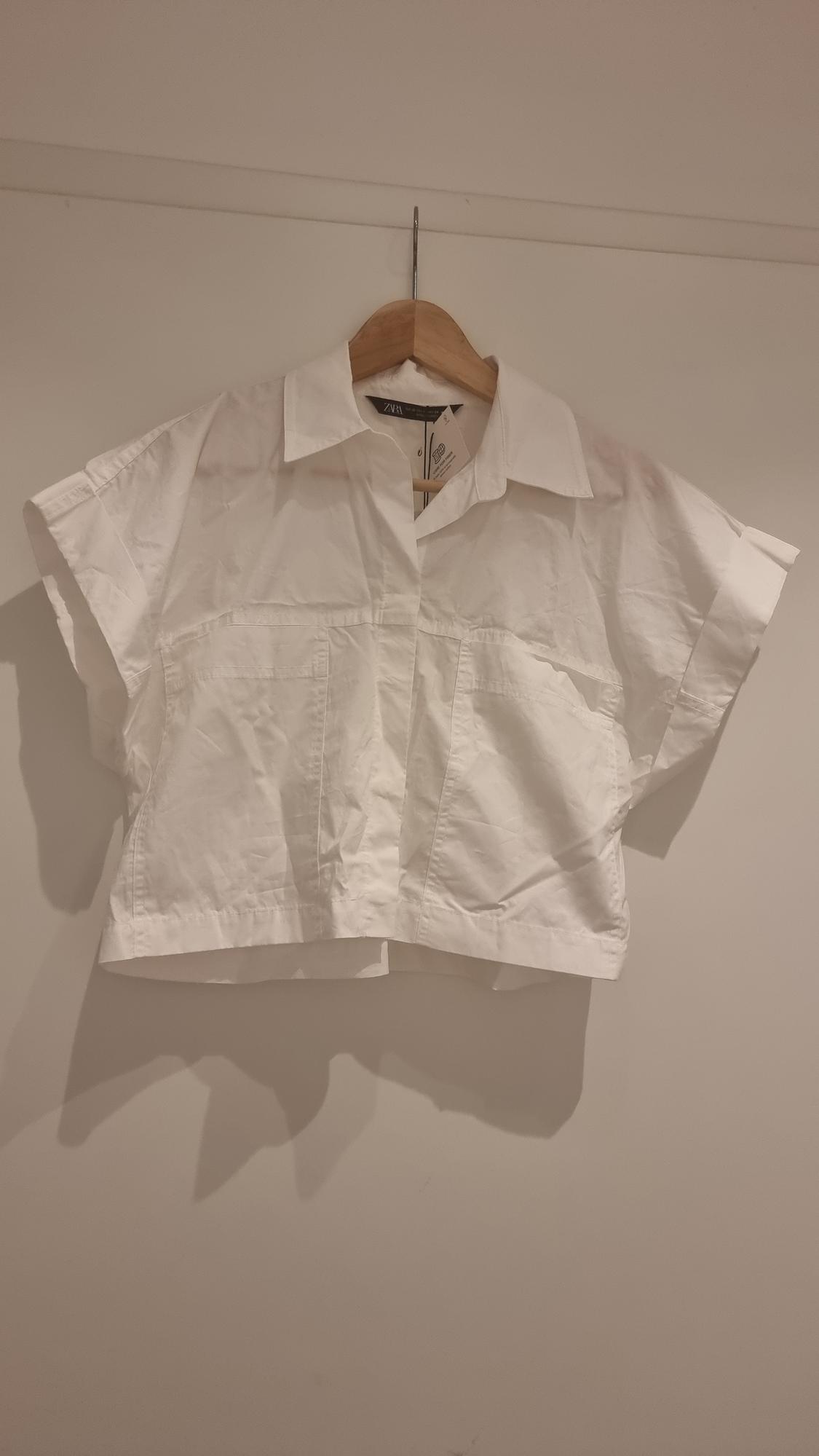 White short-sleeved shirt hanging on a wooden hanger against a white wall. Label visible on the collar.