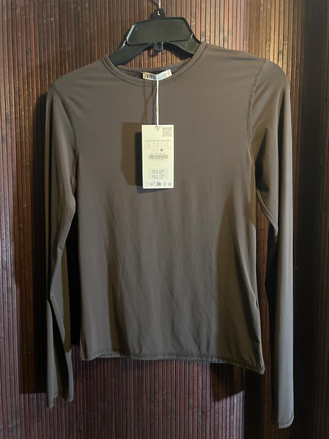Brown long-sleeve T-shirt on black hanger, with hanging price tag indicating US$ 25.95, UK£ 15.99. Wooden background.