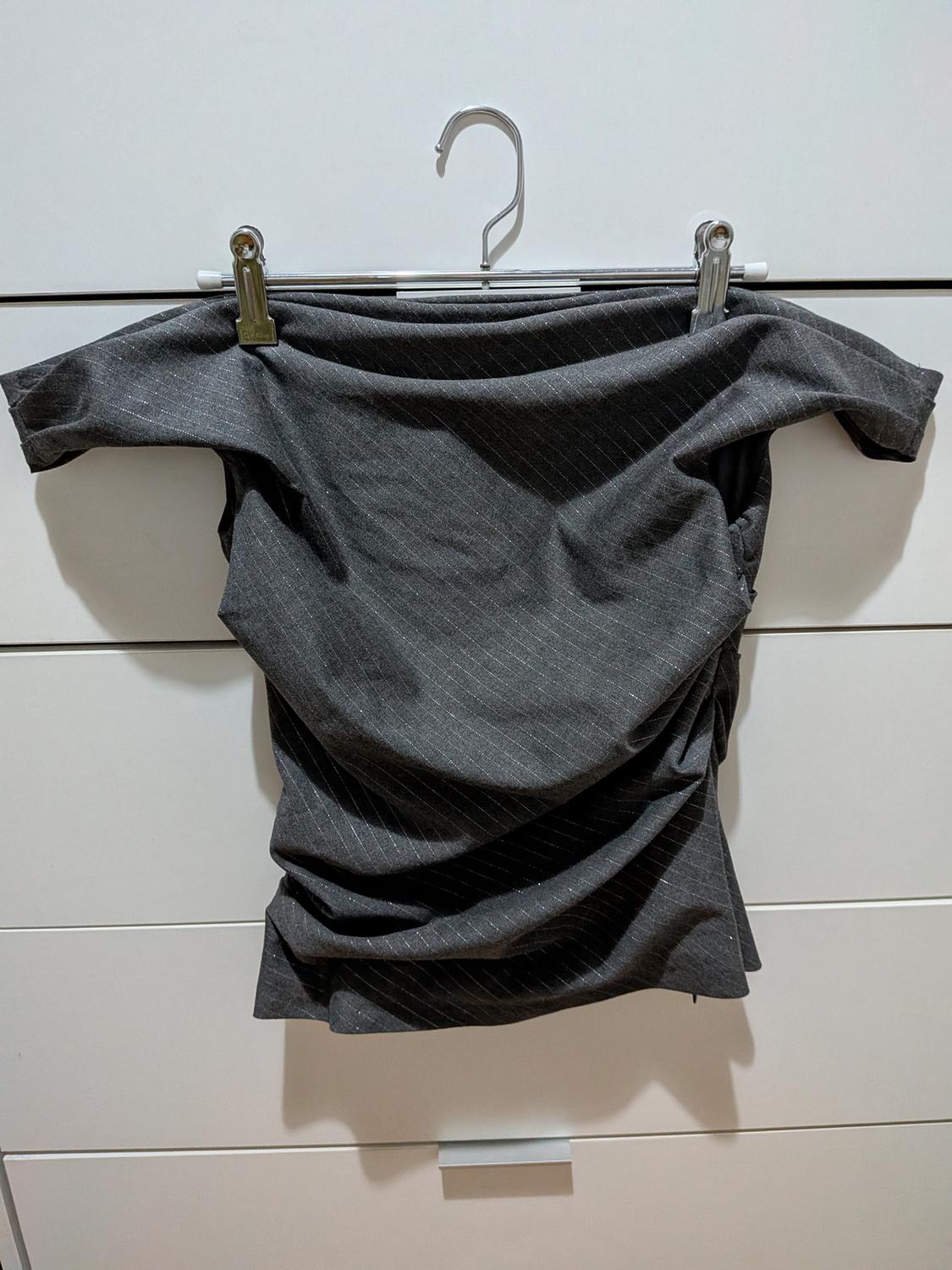 Dark grey off-the-shoulder draped top with thin light stripes, hanging on a metal hanger above white drawers.