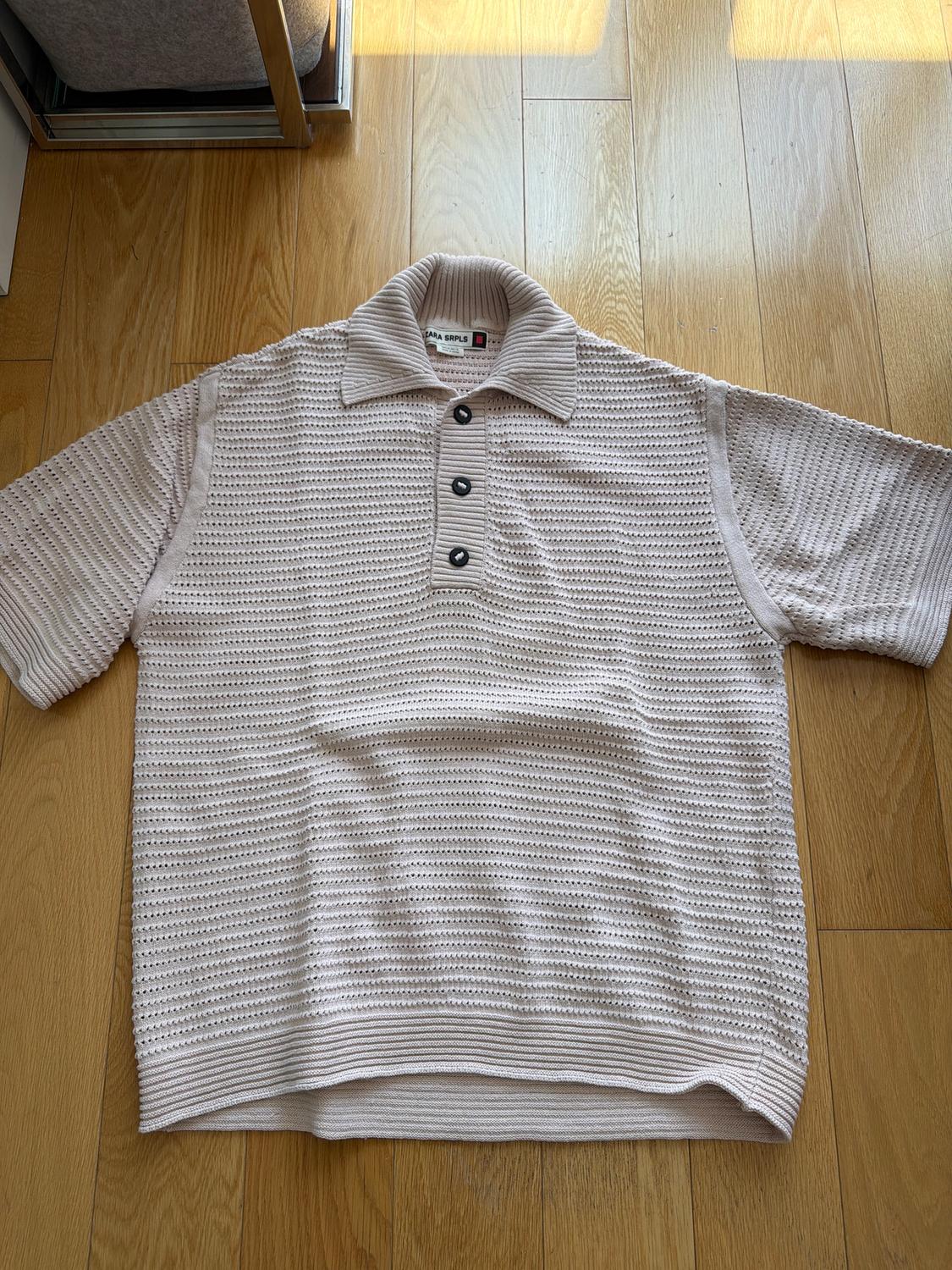 Beige knit polo with ribbed texture and three dark buttons, on a light wooden floor. Visible tag on the collar.