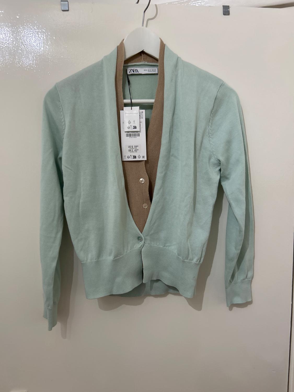 Mint green cardigan with brown collar, hanging on a white hanger. Visible label with size and brand details.