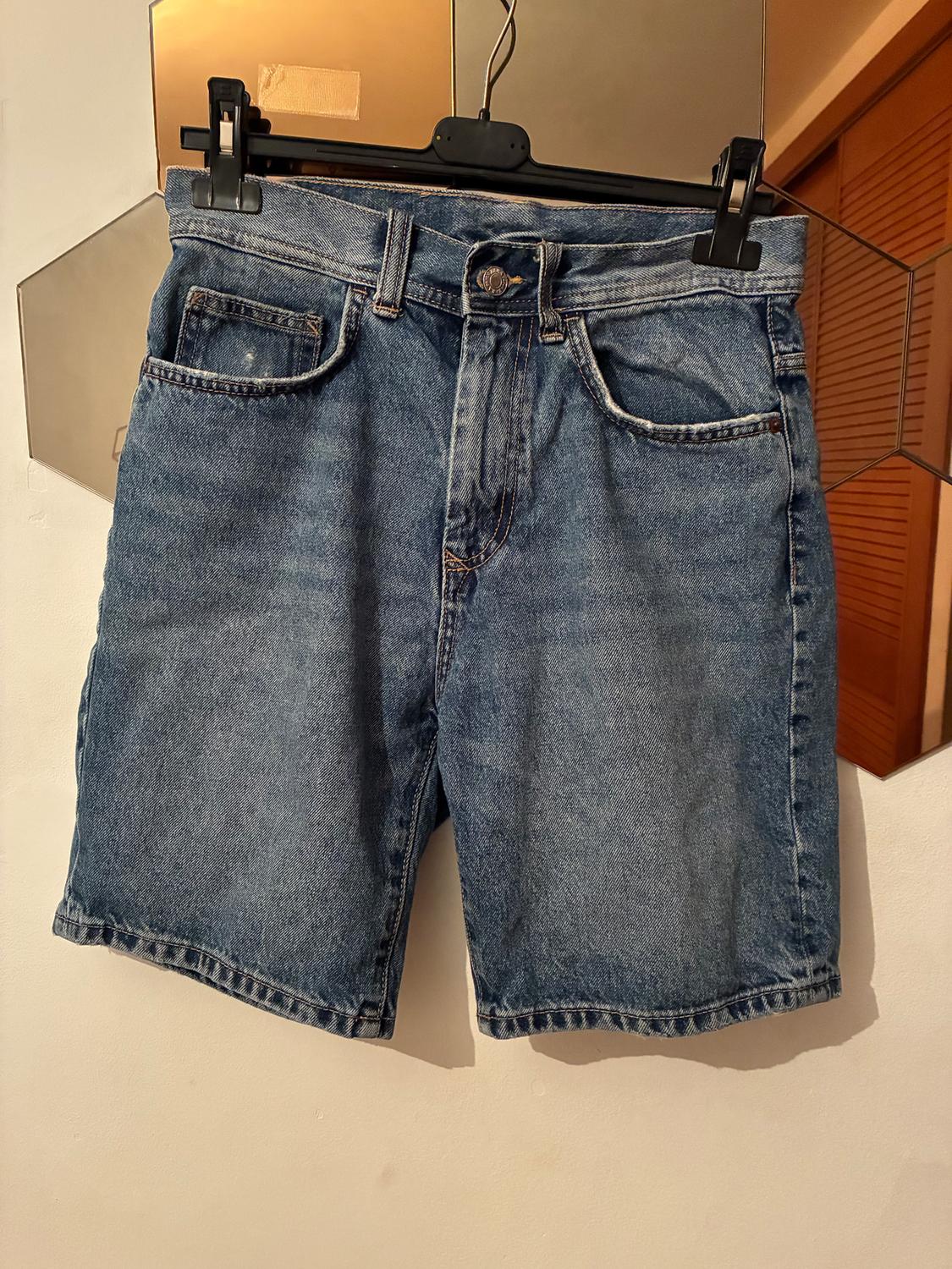 Blue denim shorts hanging on a black hanger in front of a hexagonal mirror in a well-lit room.