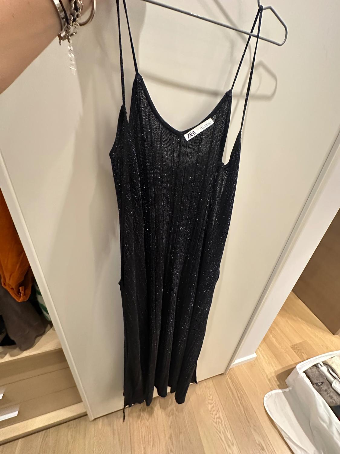 Shiny black dress hanging on a hanger, with thin straps. Visible label with the text "ZARA". Wardrobe background and wooden floor.