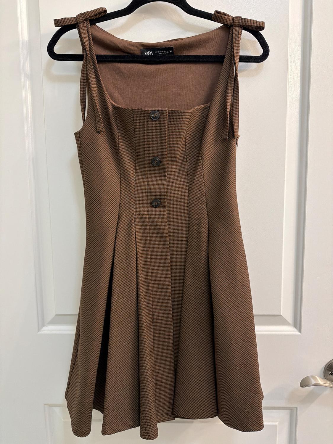 Brown dress with adjustable straps and front buttons, hanging on a black hanger over a white door.
