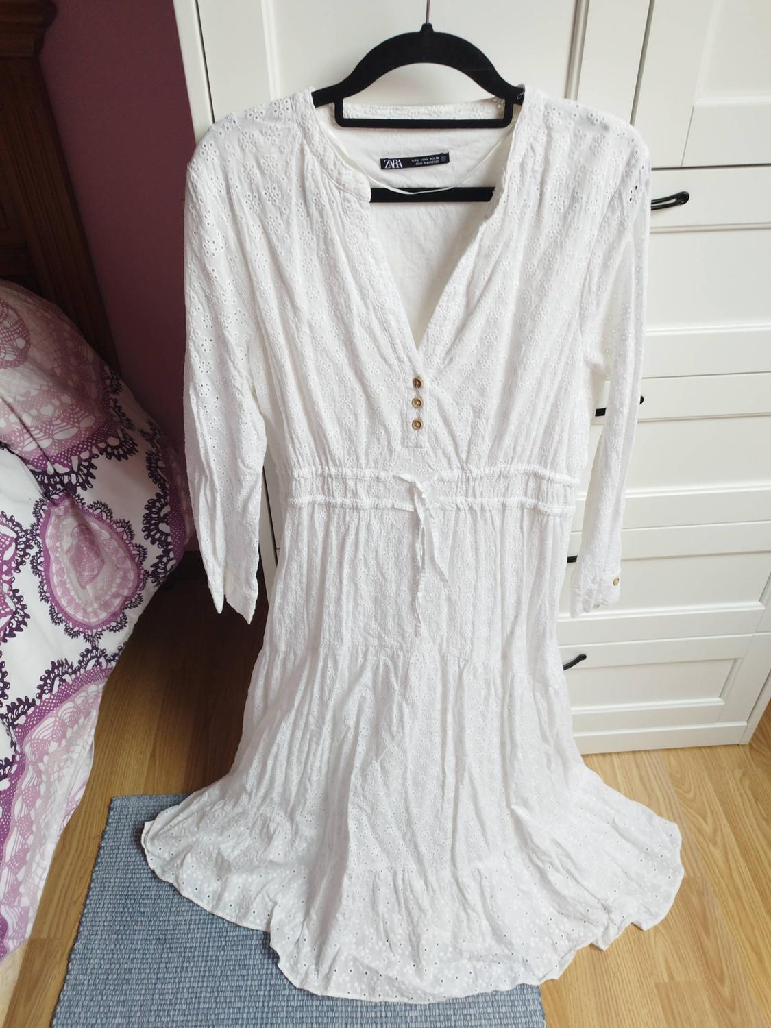 White lace dress with buttons on the chest, hanging on a black hanger in front of a white wardrobe.