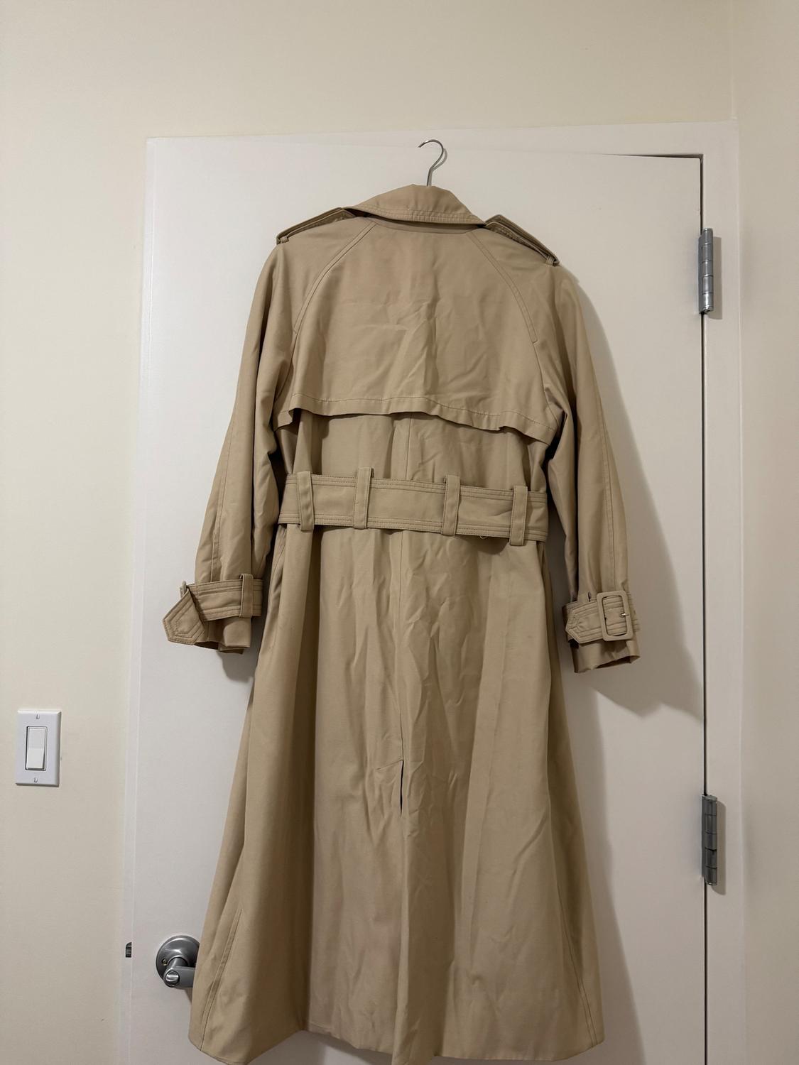 Beige trench coat hanging on a white door, seen from behind. The garment has a belt and details on the sleeves.