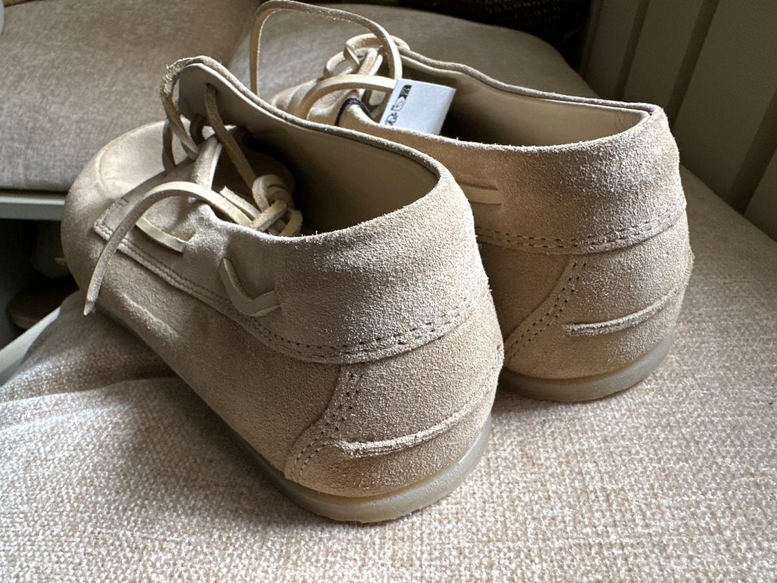 Beige suede lace-up shoes with visible stitching, seen from behind on a textured fabric surface. Relaxed atmosphere.