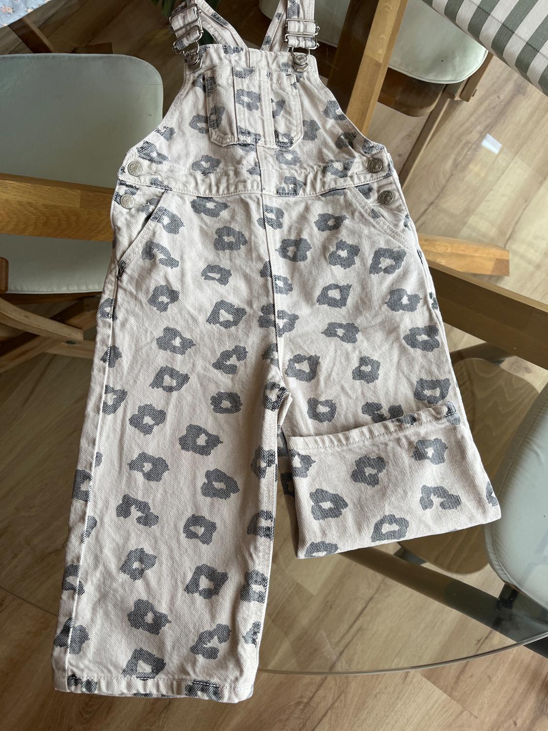 Children's beige dungarees with grey spot print on a glass table. Background of chairs and light wooden floor.