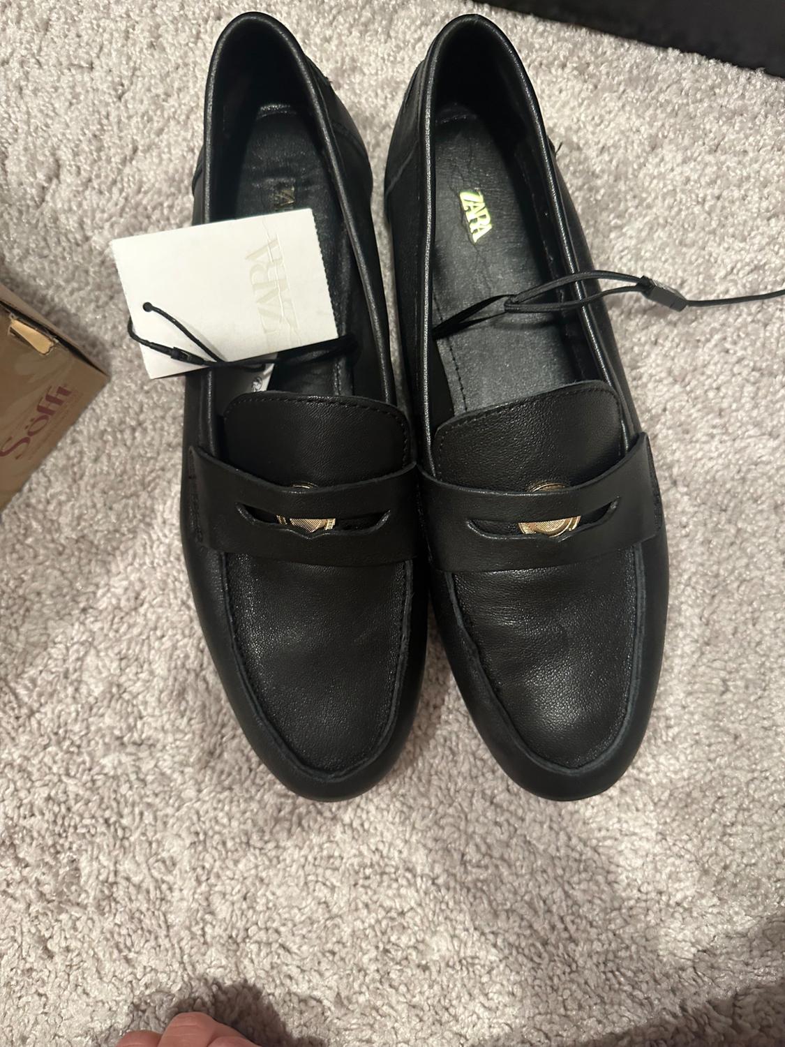 Black loafers with metallic detail on a grey rug. Includes a label with ZARA text and a side box with Söfft text.